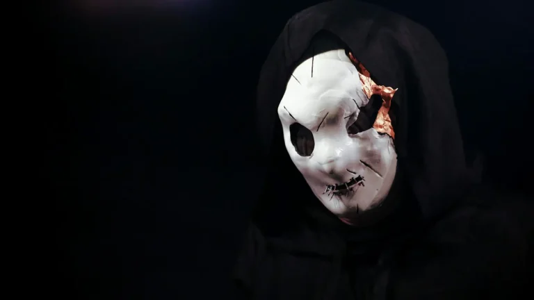 Figure in a black hood wearing a cracked white mask