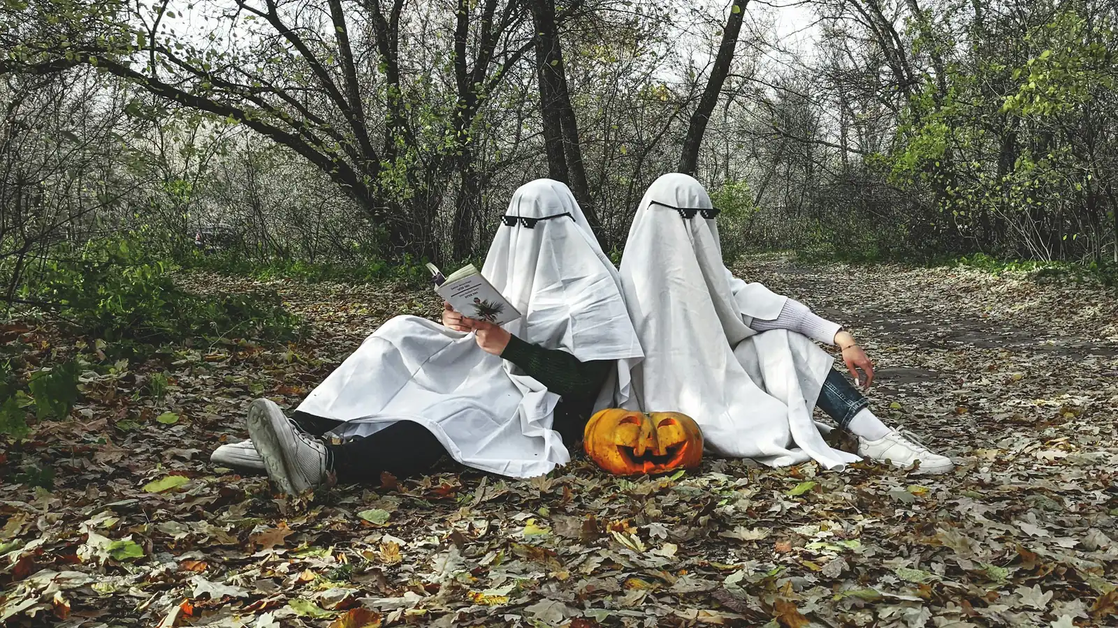 Two people in bedsheet ghost costumes sitting in autumn leaves with a pumpkin